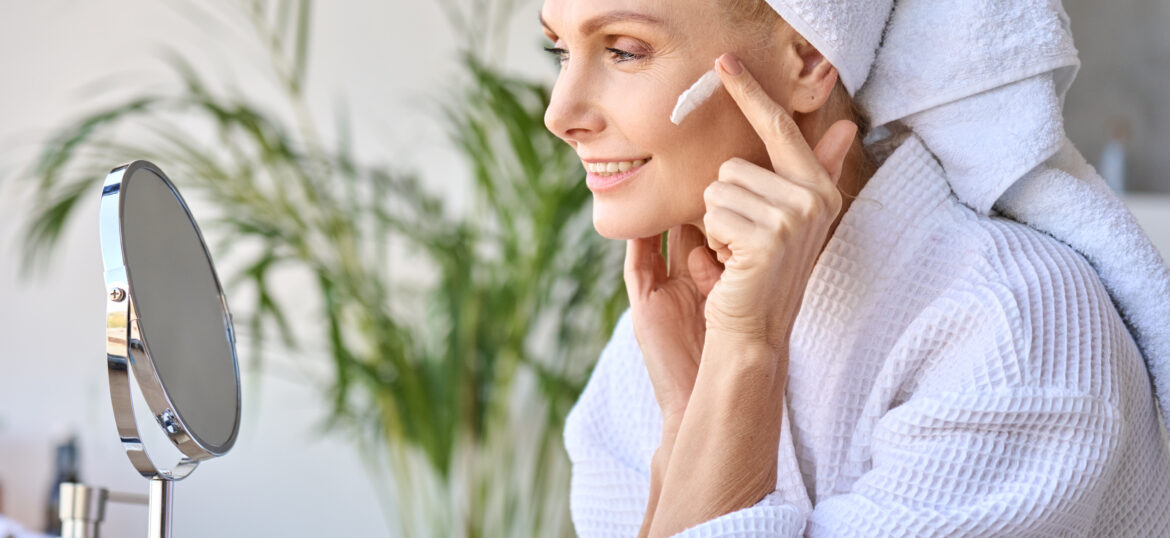 Smiling beautiful older lady looking at mirror applying cream. Skincare concept.