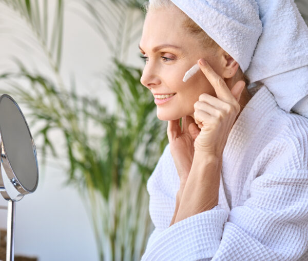 Smiling beautiful older lady looking at mirror applying cream. Skincare concept.